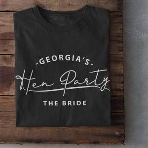 May include: Black t-shirt with white text that reads "Georgia's Hen Party The Bride".