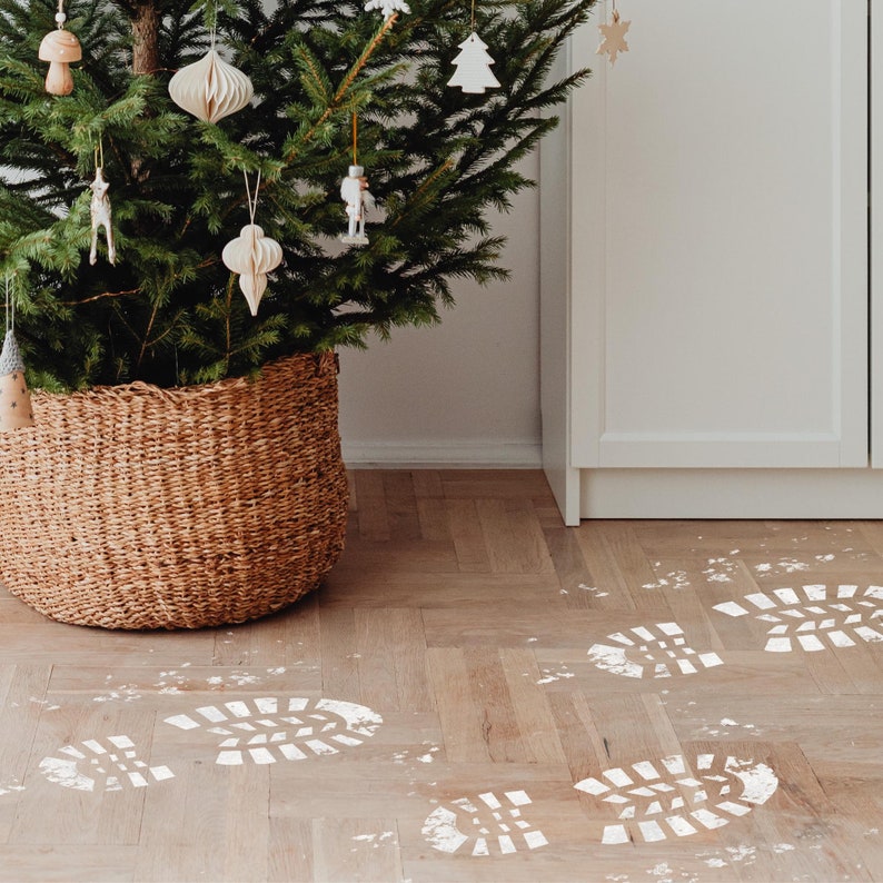 May include: A Christmas tree decorated with white ornaments sits in a wicker basket on a wood floor. White footprints are painted on the floor.