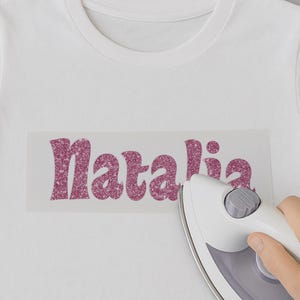 May include: A white t-shirt with the name "Natalia" in pink glitter ironed on. An iron is being used to press the glitter onto the shirt.