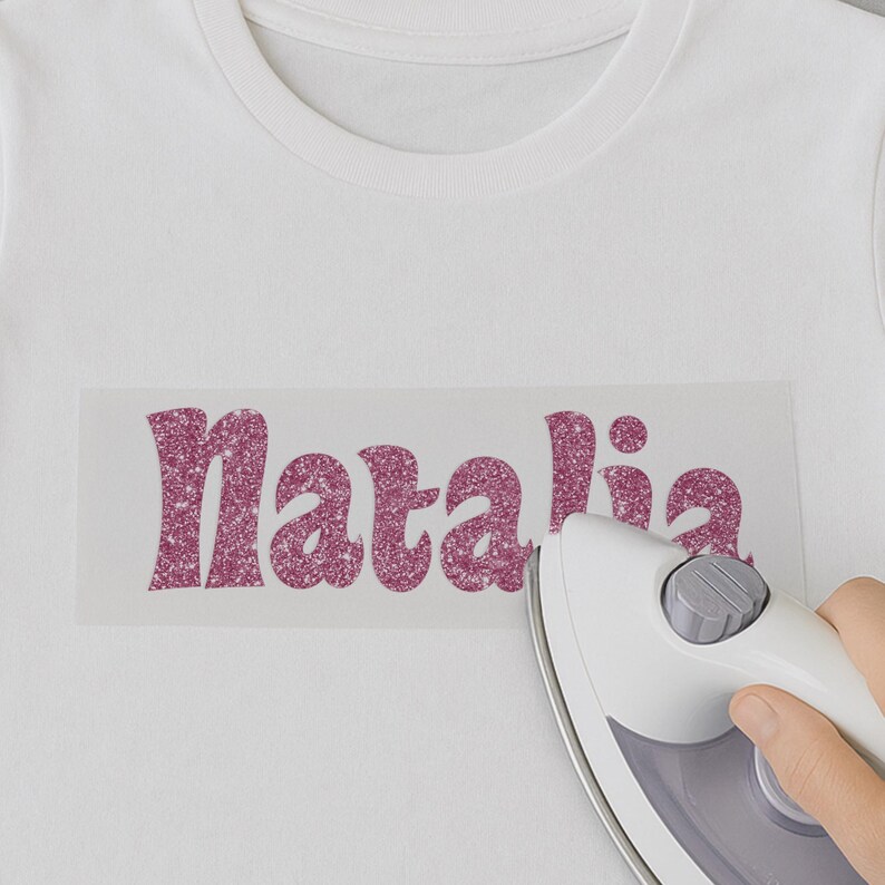 May include: A white t-shirt with the name "Natalia" in pink glitter ironed on. An iron is being used to press the glitter onto the shirt.