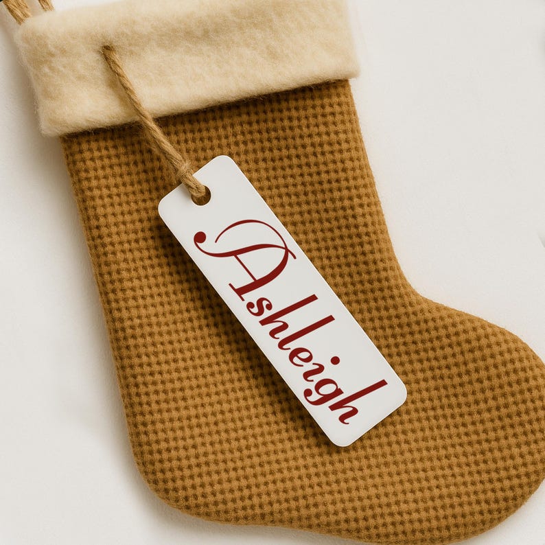 K&ouml;nnte beinhalten: Ein beigefarbener Weihnachtsstrumpf mit cremefarbenem Bund und einem Anh&auml;nger mit dem Namen "Ashleigh" in Schreibschrift. Der Strumpf besteht aus einem strukturierten Material und hat eine Schlaufe zum Aufh&auml;ngen.