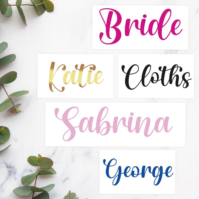 May include: Four white cards with different names written in cursive script. The names are "Bride", "Katie", "Cloths", "Sabrina", and "George". The names are written in different colors: pink, gold, black, pink, and blue.