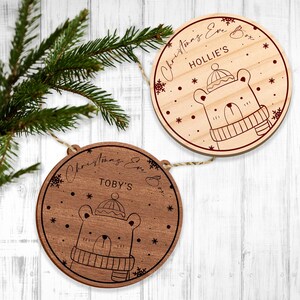 May include: Two round wooden decorations featuring a bear wearing a hat and scarf design. The decorations are adorned with snowflakes and the text "Christmas Eve Box" with a name, "Hollie's" and "Toby's".
