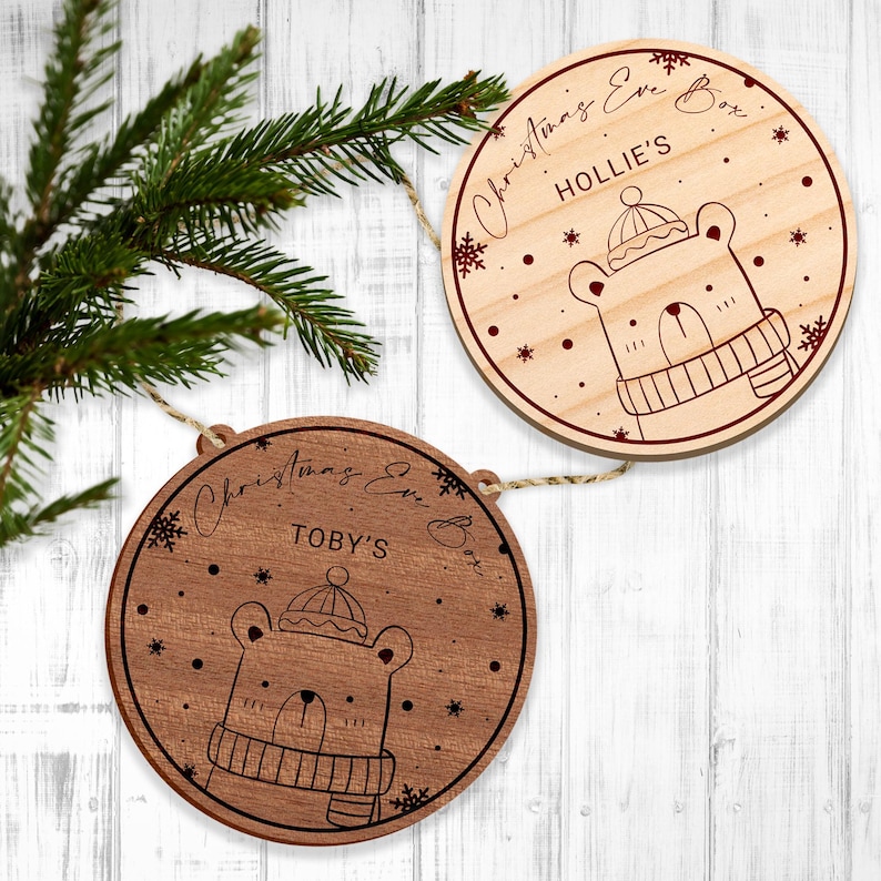 May include: Two round wooden decorations featuring a bear wearing a hat and scarf design. The decorations are adorned with snowflakes and the text "Christmas Eve Box" with a name, "Hollie's" and "Toby's".