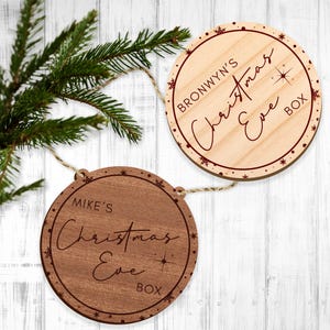 May include: Two round wooden Christmas Eve box ornaments with twine hangers. The ornament on the left is dark brown with the text "MIKE'S Christmas Eve BOX" in red. The ornament on the right is light brown with the text "BRONWYN'S Christmas Eve BOX" in red.