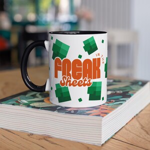 May include: White ceramic coffee mug with a black rim and handle. The mug features a green and orange design with the text "Freak Sheets" and "In The" in a retro font.