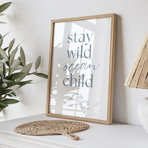 K&ouml;nnte beinhalten: Ein gerahmter Druck mit dem Text "stay wild ocean child" in blauer Schrift. Der Druck zeigt ein stilisiertes Wellendesign und ein dezentes Palmblatt-Muster im Hintergrund.