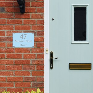 May include: A light blue house number sign with the number 47, the words "Mouse Close Drive" and silver trim. The sign is mounted on a brick wall next to a light blue door with a brass door handle and a brass mailbox.