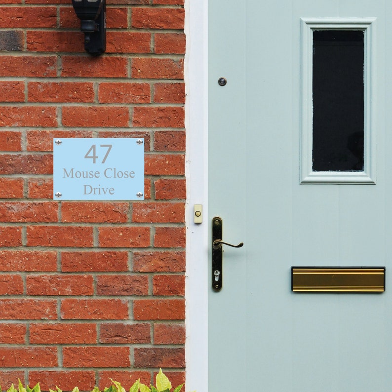 May include: A light blue house number sign with the number 47, the words "Mouse Close Drive" and silver trim. The sign is mounted on a brick wall next to a light blue door with a brass door handle and a brass mailbox.