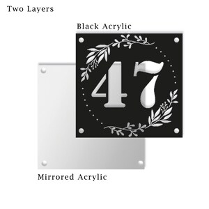May include: Two-layer house number sign with the number 47 in white on a black background. The number is surrounded by a white floral wreath. The sign is made of acrylic.