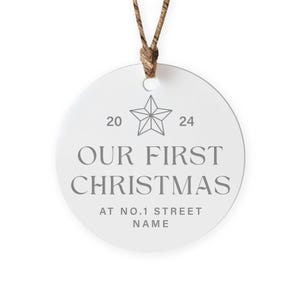 May include: White round Christmas ornament with a gray star and the text "Our First Christmas at No. 1 Street Name" with the numbers 20 and 24 on either side of the star.