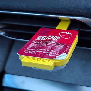 May include: A yellow plastic car air freshener shaped like a ketchup packet. The packet has a red tomato image and the text "KETCHUP 21ml e Best Before: See Pot" printed on it. The air freshener has the word "SAUCE" printed on the bottom.