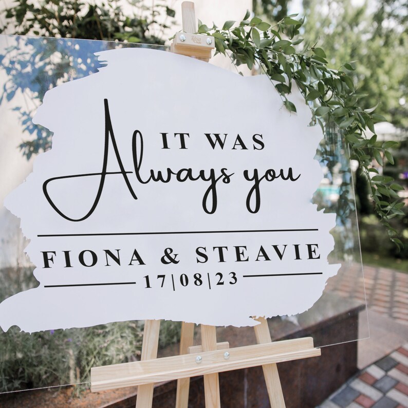 May include: A white acrylic sign with black calligraphy that reads "It was always you" with the names Fiona & Steavie and the date 17/08/23 below. The sign is propped up on a wooden easel.