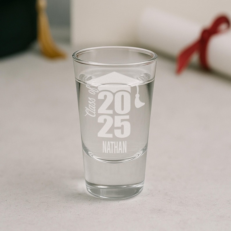 May include: A clear shot glass filled with liquid, featuring the text "Class of 2025" and the name "Nathan" in white. A graduation cap graphic is also visible. The glass is on a light surface.