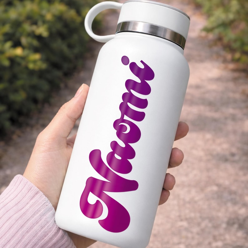 May include: A white water bottle with a silver rim and handle. The bottle features the name "Naomi" in a cursive, magenta font. The bottle is held in a person's hand.