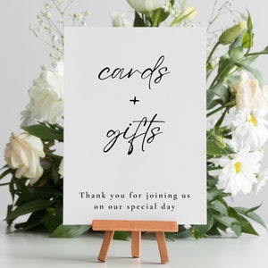 May include: A white sign on a small wooden easel with the words "cards + gifts" in elegant black script. The sign also includes the text "Thank you for joining us on our special day." White flowers and greenery frame the sign.