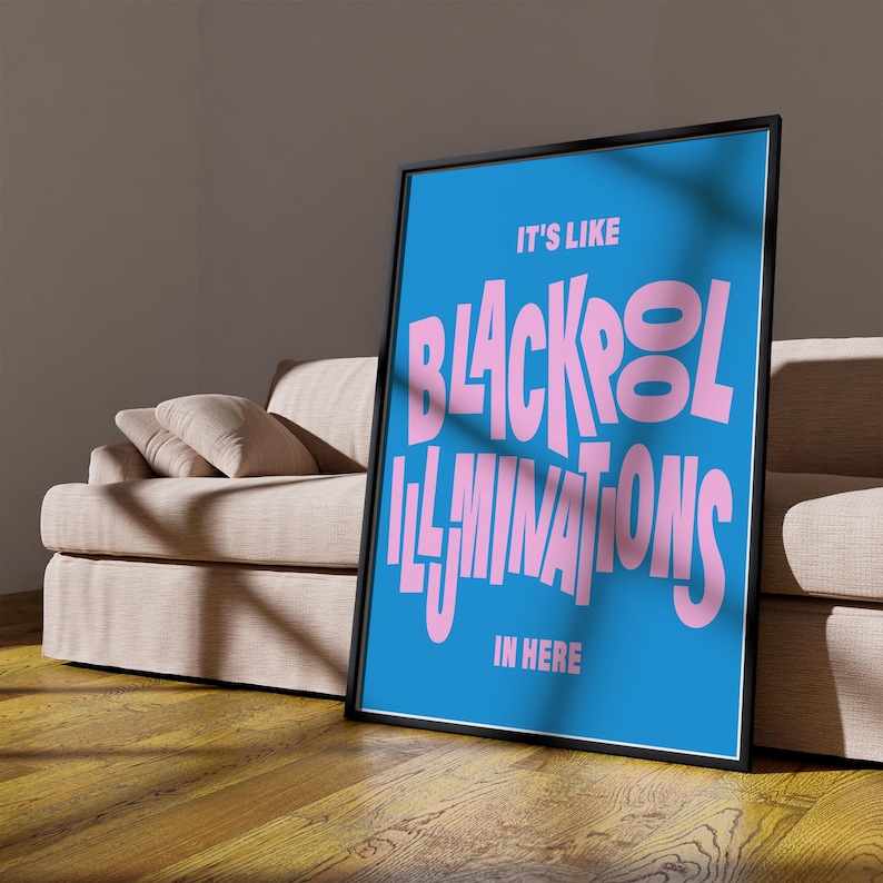 May include: A framed poster with a blue background and pink text that reads "IT'S LIKE BLACKPOOL ILLUMINATIONS IN HERE." The poster is leaning against a beige sofa and is displayed on a wooden floor.