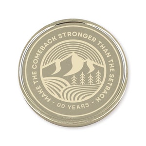 May include: A silver-colored coin with a white design. The coin features a mountain range, trees, and the text "Make the comeback stronger than the setback - 00 years - "
