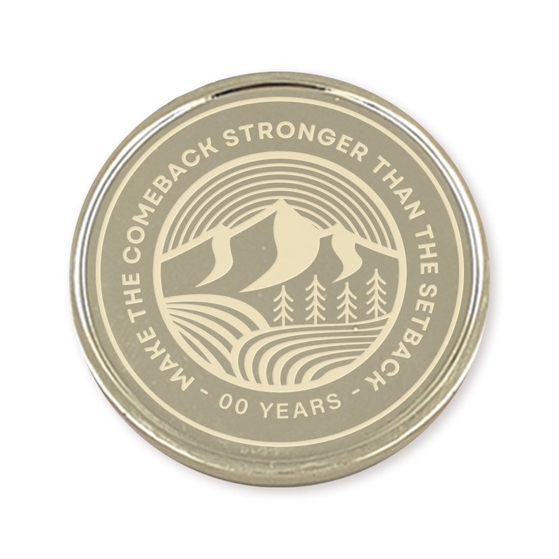 May include: A silver-colored coin with a white design. The coin features a mountain range, trees, and the text "Make the comeback stronger than the setback - 00 years - "