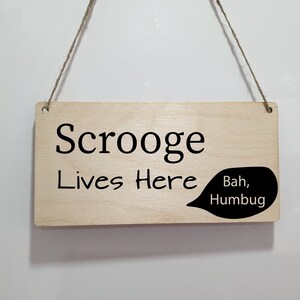 May include: A wooden sign with the text "Scrooge Lives Here" and a speech bubble that says "Bah, Humbug".