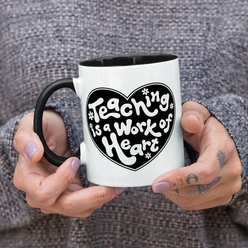 Puede incluir: Taza de caf&eacute; blanca con asa negra y borde negro. La taza tiene un coraz&oacute;n negro con el texto "Teaching is a work of heart" en letras blancas.