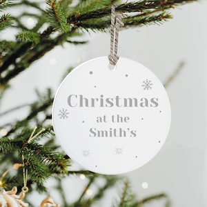 Peut inclure: Une d&eacute;coration de No&euml;l ronde et blanche avec le texte "Christmas at the Smith's" en gris. L'ornement est orn&eacute; de flocons de neige et de points. Il est suspendu par une ficelle en ficelle naturelle, sur un sapin de No&euml;l.