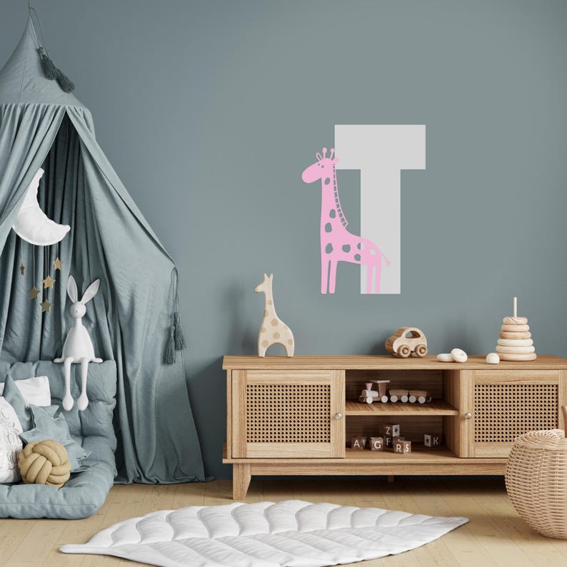 May include: A pink giraffe with black spots stands next to a white letter T on a light blue wall. A wooden shelf with a wicker basket and a toy car sits below the wall decal.