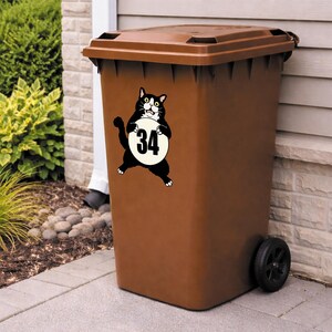 May include: A brown outdoor trash bin with a black cat sticker. The cat is holding a white circle with the number 34 in black. The bin has a brown lid and a black wheel.