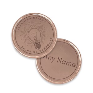 Puede incluir: Dos fichas redondas de color oro rosa. Una ficha presenta un dise&ntilde;o de bombilla grabado con el texto "TEACHING BRIGHT STUDENTS TO SHINE". La otra ficha tiene el texto grabado "Any Name".