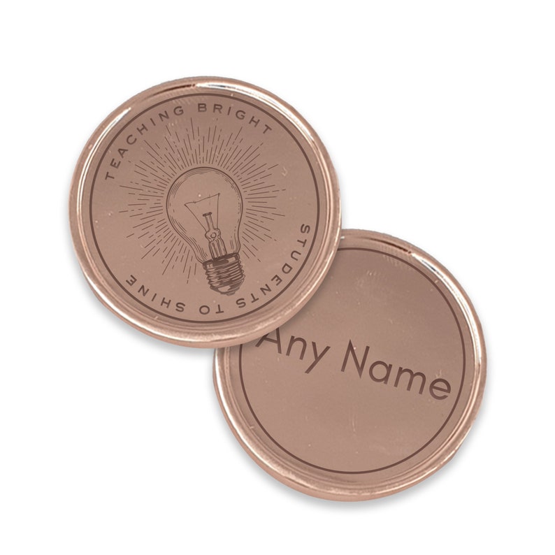 Puede incluir: Dos fichas redondas de color oro rosa. Una ficha presenta un dise&ntilde;o de bombilla grabado con el texto "TEACHING BRIGHT STUDENTS TO SHINE". La otra ficha tiene el texto grabado "Any Name".