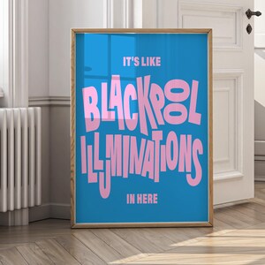 May include: A framed poster with a blue background and pink text. The text reads "IT'S LIKE BLACKPOOL ILLUMINATIONS IN HERE". The frame is a light brown color. The poster is displayed in a room with a white door and wooden floor.