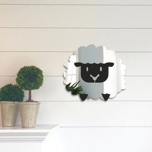 May include: A sheep-shaped wall mirror with a black face and feet, and a white, fluffy outline. The mirror is mounted on a white wall with horizontal paneling. Two potted topiary plants are on a shelf below.