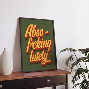 Puede incluir: Una impresi&oacute;n art&iacute;stica enmarcada con las palabras "Abso-f*cking-lutely" en letras amarillas y rojas sobre un fondo verde oscuro. La impresi&oacute;n se muestra sobre una mesa marr&oacute;n contra una pared blanca.