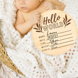 K&ouml;nnte beinhalten: Eine runde Holzplakette mit dem Text "Hello World" und einem floralen Design. Die Plakette hat Platz zum Ausf&uuml;llen des Namens des Babys, des Datums, der Uhrzeit, des Gewichts und der L&auml;nge. Der Name ist "Lauren", das Datum ist "09/07/23", die Uhrzeit ist "9:36 Uhr", das Gewicht ist "9 lbs" und die L&auml;nge ist "21 in".