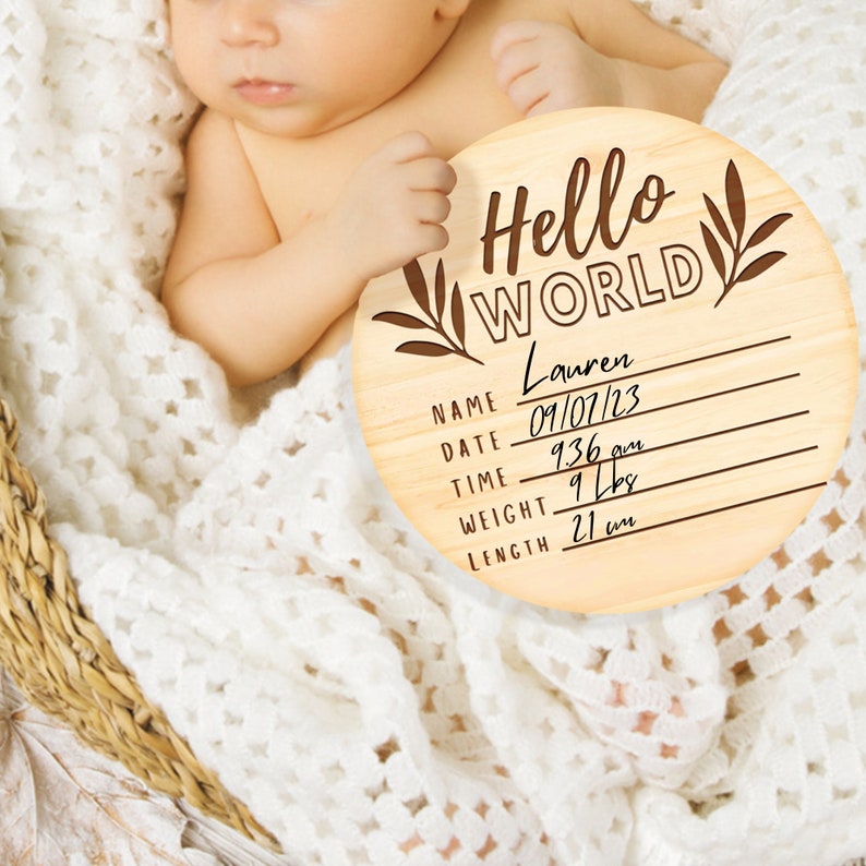 K&ouml;nnte beinhalten: Eine runde Holzplakette mit dem Text "Hello World" und einem floralen Design. Die Plakette hat Platz zum Ausf&uuml;llen des Namens des Babys, des Datums, der Uhrzeit, des Gewichts und der L&auml;nge. Der Name ist "Lauren", das Datum ist "09/07/23", die Uhrzeit ist "9:36 Uhr", das Gewicht ist "9 lbs" und die L&auml;nge ist "21 in".