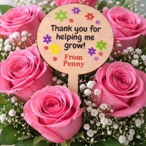May include: A bouquet of pink roses with white baby's breath and green leaves. A wooden sign with the text "Thank you for helping me grow! From Penny" is placed in the center of the arrangement. The sign is decorated with colorful flower accents.
