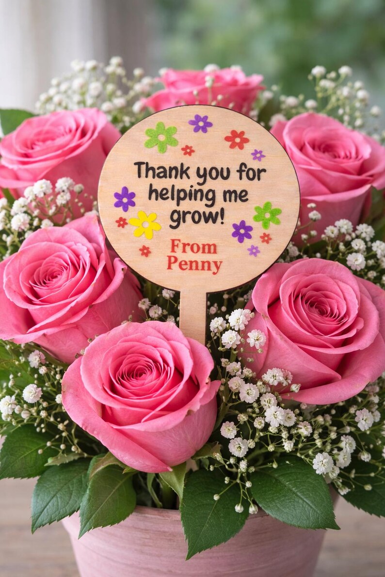 May include: A bouquet of pink roses with white baby's breath and green leaves. A wooden sign with the text "Thank you for helping me grow! From Penny" is placed in the center of the arrangement. The sign is decorated with colorful flower accents.