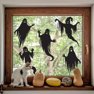 May include: Five black ghost silhouettes are displayed on a window with a cat sitting on the windowsill. The windowsill has several gourds and squash in front of the cat.