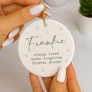 May include: A round, off-white ceramic ornament with the name "Frankie" in a script font. The ornament features paw prints and the text "always loved never forgotten forever missed" in a muted green color. A jute twine loop is attached for hanging.