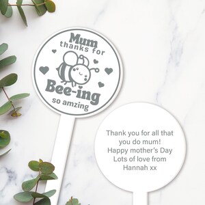 K&ouml;nnte beinhalten: Ein wei&szlig;er Gartenpfahl mit einer grauen Bienenillustration und dem Text "Mum thanks for Bee-ing so amzing". Ein zweiter wei&szlig;er Pfahl tr&auml;gt den Text "Thank you for all that you do mum! Happy mother's Day Lots of love from Hannah xx".