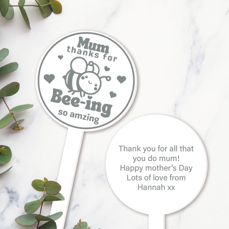 K&ouml;nnte beinhalten: Ein wei&szlig;er Gartenpfahl mit einer grauen Bienenillustration und dem Text "Mum thanks for Bee-ing so amzing". Ein zweiter wei&szlig;er Pfahl tr&auml;gt den Text "Thank you for all that you do mum! Happy mother's Day Lots of love from Hannah xx".