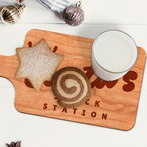 Op de afbeelding: Houten serveerplank met een stervormige koek, een swirlkoekje en een glas melk. Op de plank staat de tekst "Santa's Snack Station". Kerstversieringen zijn op de achtergrond.