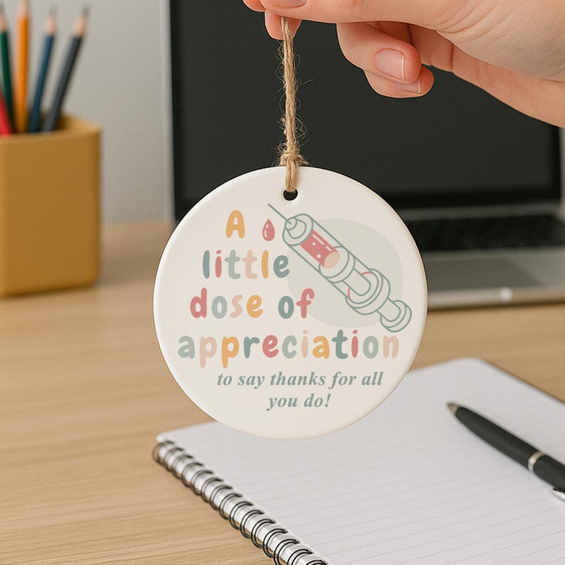 May include: A round, white ceramic ornament with the text "A little dose of appreciation to say thanks for all you do!" and a syringe illustration. The ornament is suspended by a natural twine string.