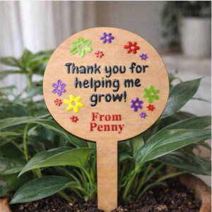 May include: A wooden plant stake with a round top and a rectangular base. The top has the text "Thank you for helping me grow!" and "From Penny" in black and red. Colorful flower accents surround the text. The stake is placed in a potted plant.