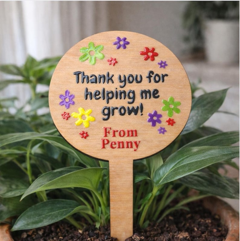 May include: A wooden plant stake with a round top and a rectangular base. The top has the text "Thank you for helping me grow!" and "From Penny" in black and red. Colorful flower accents surround the text. The stake is placed in a potted plant.