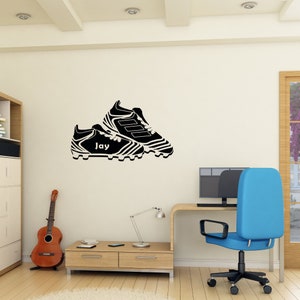 May include: Black and white silhouette of a pair of soccer cleats with the name "Jay" written on the side. The image is on a white wall in a room with a desk, chair, and guitar.