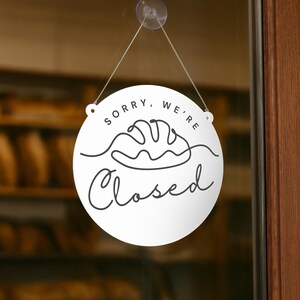 以下が含まれることがあります： 「SORRY, WE'RE Closed」の黒い筆記体で書かれた白い丸い看板。パンの連続線画が「Closed」という単語の上にあります。看板はガラスのドアに吊り下げられています。