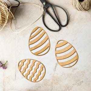 May include: Three wooden Easter egg shapes with different patterns. The eggs are cut out of wood and have a natural brown colour. The patterns are stripes, waves, and a more intricate design.