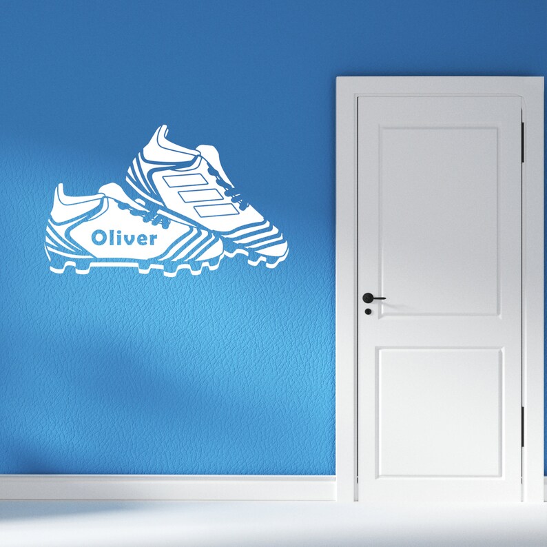 May include: White vinyl decal of a pair of soccer cleats with the name "Oliver" on one cleat. The decal is on a blue wall.