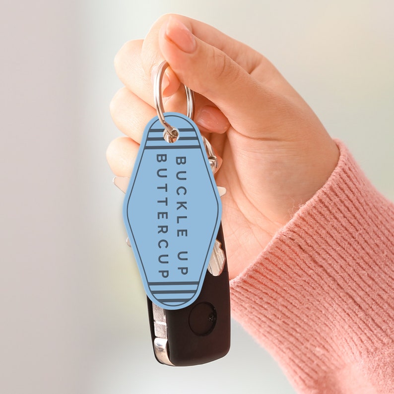Puede incluir: Un llavero azul en forma de llave de motel con el texto "buckle up buttercup" en letras negras. El llavero est&aacute; unido a un llavero plateado y a una llave de coche negra.